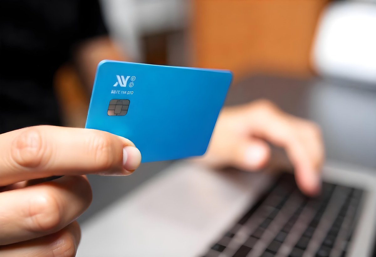 A Brown Skin Hand Holding A Blue Credit Card Over A Laptop The Background Is Blurred And Looks Like An Office 1 E660