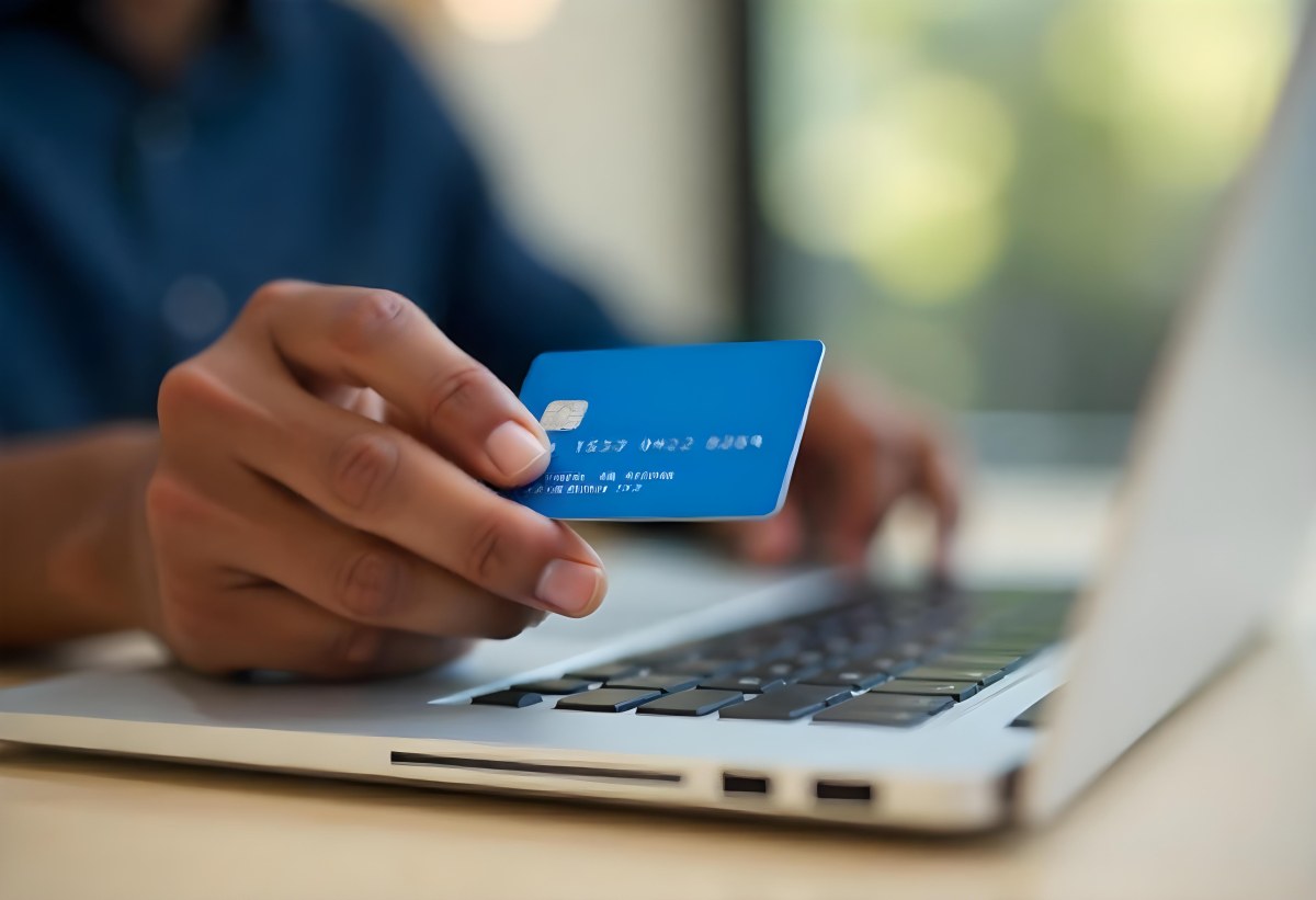 A Brown Skin Hand Holding A Blue Credit Card Over A Laptop The Background Is Blurred And Looks Like An Office 10 3d37