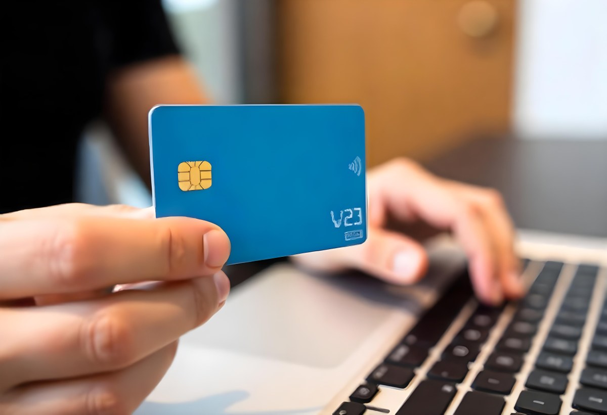 A Brown Skin Hand Holding A Blue Credit Card Over A Laptop The Background Is Blurred And Looks Like An Office 11 3ab1