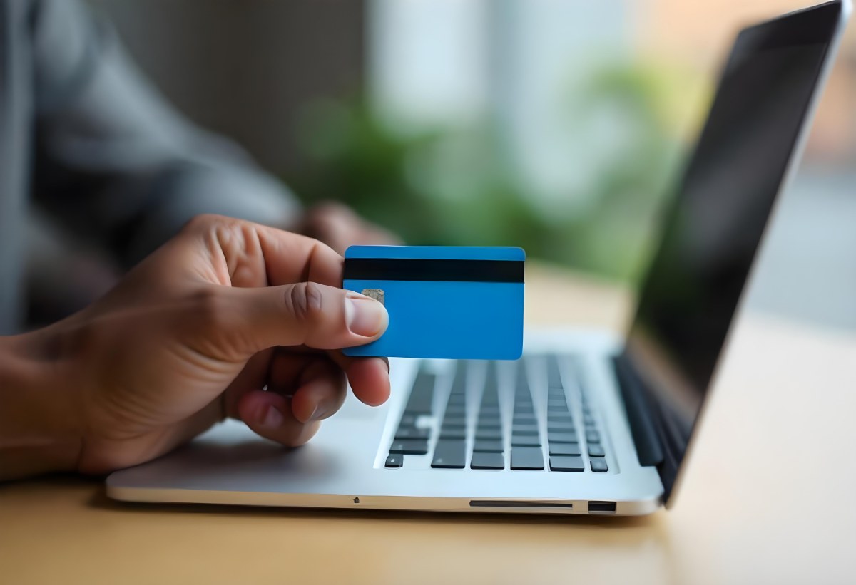 A Brown Skin Hand Holding A Blue Credit Card Over A Laptop The Background Is Blurred And Looks Like An Office 13 1b91