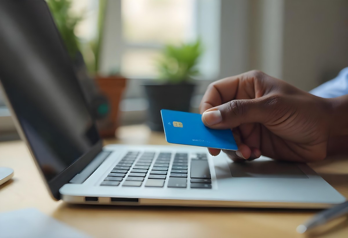 A Brown Skin Hand Holding A Blue Credit Card Over A Laptop The Background Is Blurred And Looks Like An Office 16 A231