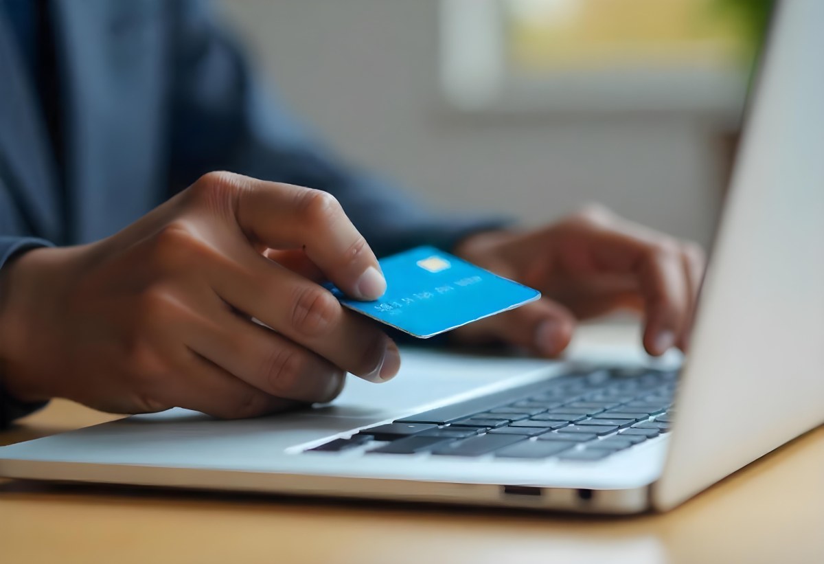 A Brown Skin Hand Holding A Blue Credit Card Over A Laptop The Background Is Blurred And Looks Like An Office 17 78c2