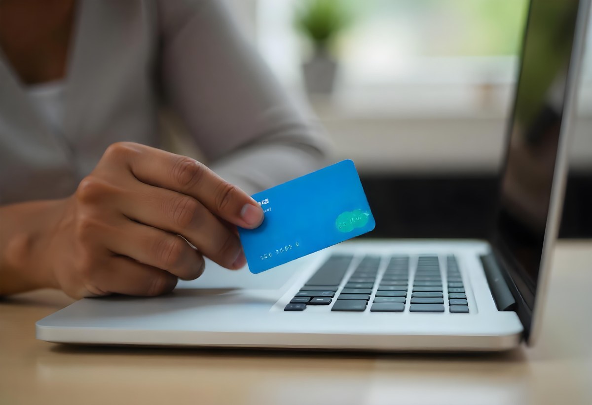 A Brown Skin Hand Holding A Blue Credit Card Over A Laptop The Background Is Blurred And Looks Like An Office 18 6a6d