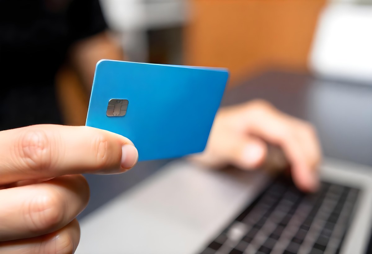A Brown Skin Hand Holding A Blue Credit Card Over A Laptop The Background Is Blurred And Looks Like An Office 2 Ac60