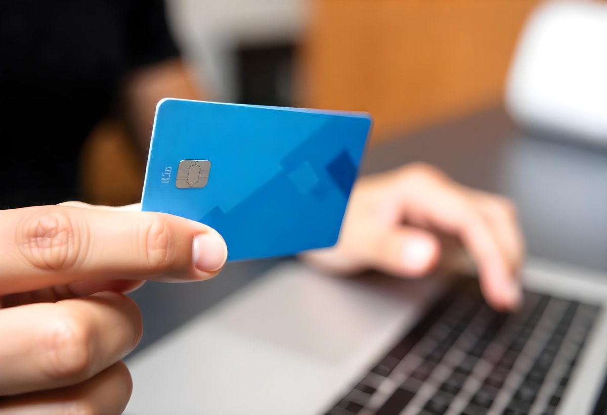 A Brown Skin Hand Holding A Blue Credit Card Over A Laptop The Background Is Blurred And Looks Like An Office 3 A88f