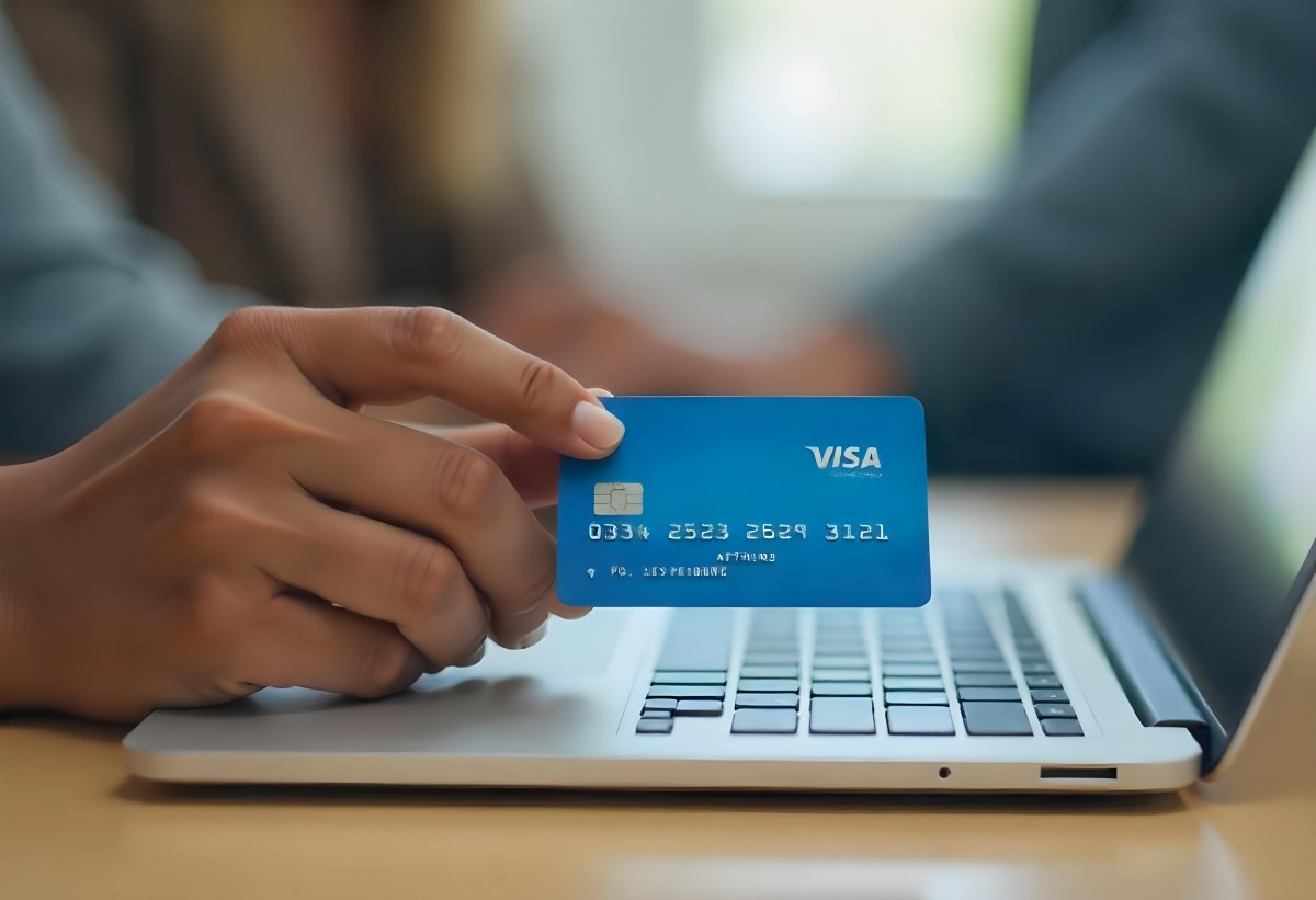 A Brown Skin Hand Holding A Blue Credit Card Over A Laptop The Background Is Blurred And Looks Like An Office 5 A2d3