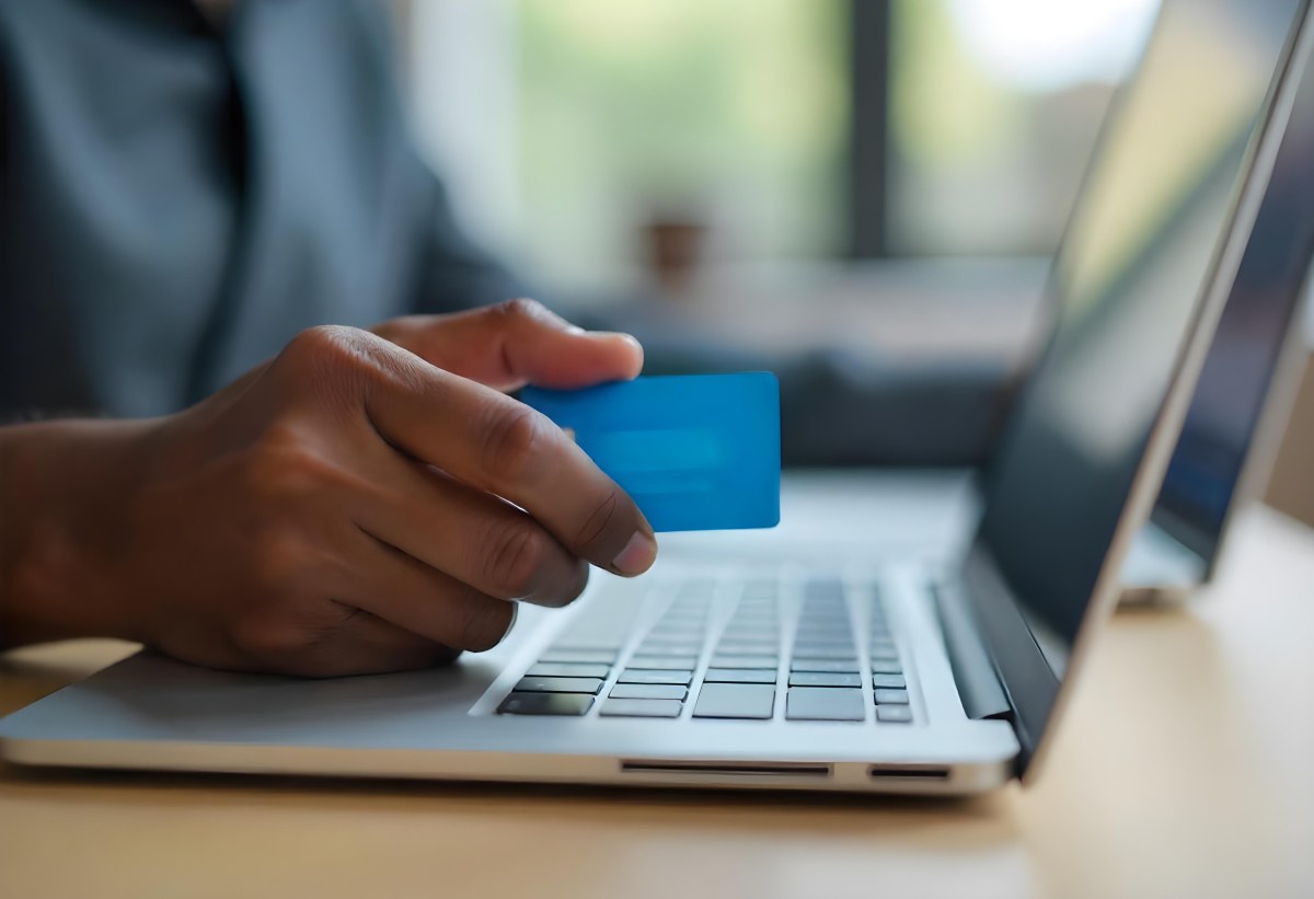 A Brown Skin Hand Holding A Blue Credit Card Over A Laptop The Background Is Blurred And Looks Like An Office 6 7dc1