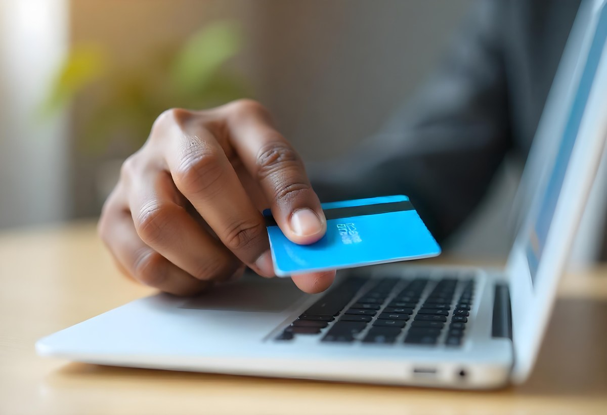 A Brown Skin Hand Holding A Blue Credit Card Over A Laptop The Background Is Blurred And Looks Like An Office 7 A2b3