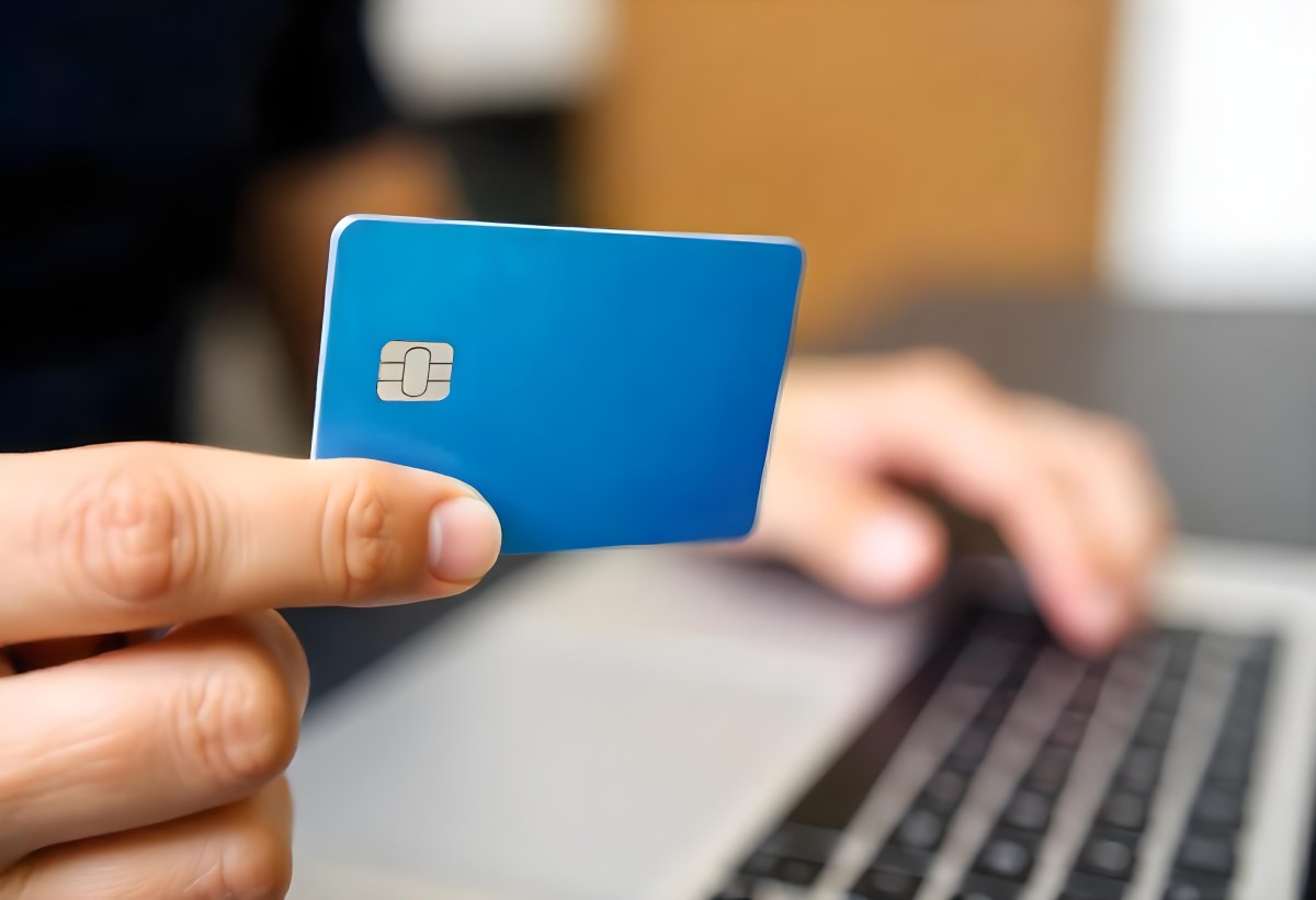 A Brown Skin Hand Holding A Blue Credit Card Over A Laptop The Background Is Blurred And Looks Like An Office 8 9f23