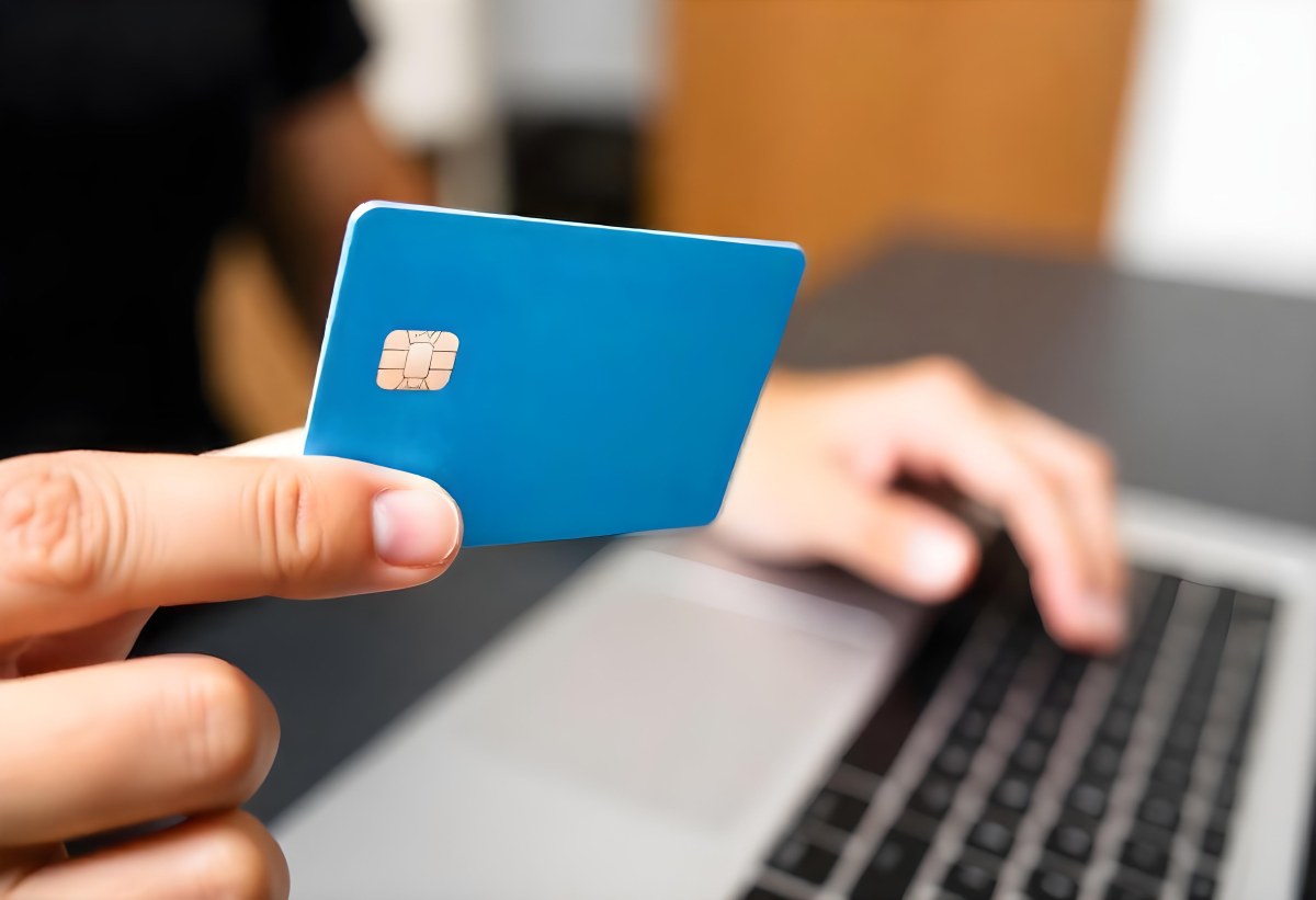 A Brown Skin Hand Holding A Blue Credit Card Over A Laptop The Background Is Blurred And Looks Like An Office 9 C77e