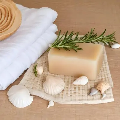 A Bar Of Soap Placed On A Woven Mat Surrounded By Various Seashells And A Sprig Of Rosemary In The Background There Are Neatly Stacked White Towels And A Natural Sponge The Setting Has A Calming Spa Like Atmospher 25 81b5