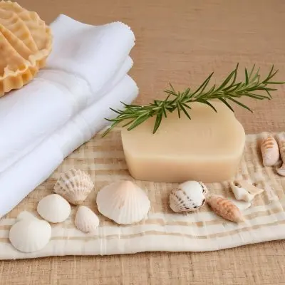 A Bar Of Soap Placed On A Woven Mat Surrounded By Various Seashells And A Sprig Of Rosemary In The Background There Are Neatly Stacked White Towels And A Natural Sponge The Setting Has A Calming Spa Like Atmospher 26 0dc3
