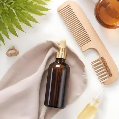 A Bottle Of Amber Colored Hair Oil Product Surrounded By Green Leaves And Hair Styling Tools On A White Background 9