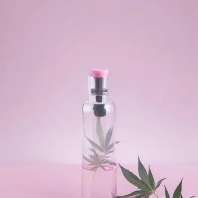 A Clear Glass Bottle With A Pink Cap Placed On A Pink Surface Cannabis Leaves In The Foreground Light Purple Background 1 5953