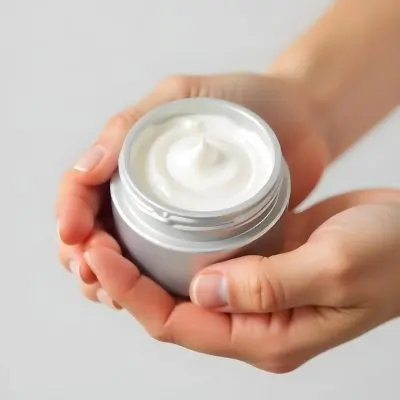 A Pair Of Fair Skinned Hands Holding A Silver Jar Of White Cream One Finger Touching The Cream Plain Background 19 528a