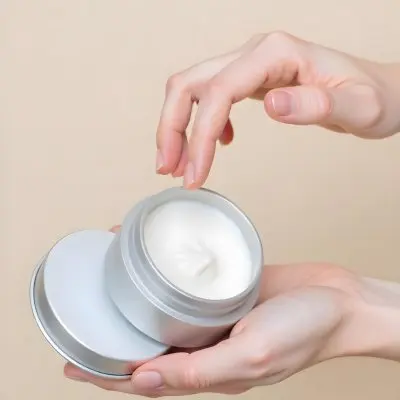 A Pair Of Fair Skinned Hands Holding A Silver Jar Of White Cream One Finger Touching The Cream Plain Background 2 E193