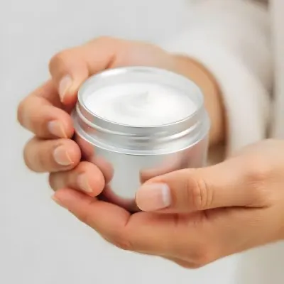 A Pair Of Fair Skinned Hands Holding A Silver Jar Of White Cream One Finger Touching The Cream Plain Background 20 20e3