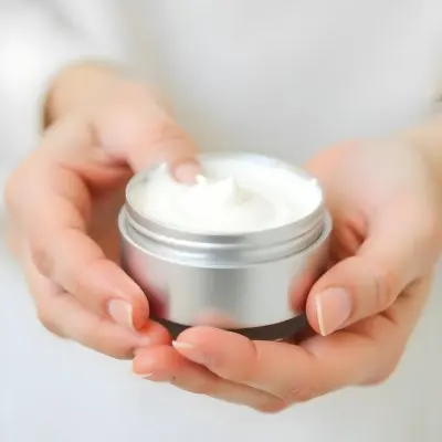 A Pair Of Fair Skinned Hands Holding A Silver Jar Of White Cream One Finger Touching The Cream Plain Background 21 Bcfb