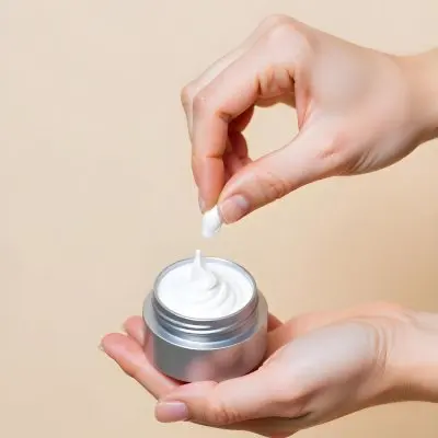 A Pair Of Fair Skinned Hands Holding A Silver Jar Of White Cream One Finger Touching The Cream Plain Background 22 Aace