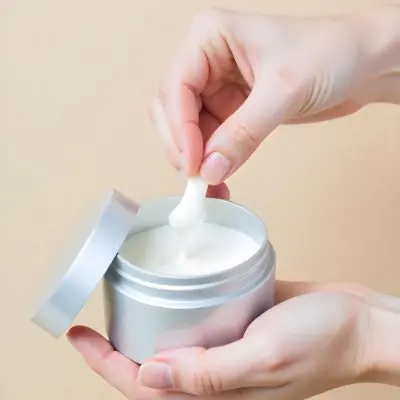 A Pair Of Fair Skinned Hands Holding A Silver Jar Of White Cream One Finger Touching The Cream Plain Background 3 8a20
