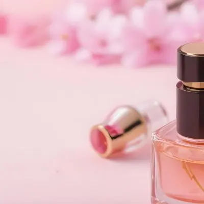 A Perfume Bottle With A Golden Cap Pink Liquid Inside Blurred Pink Flowers In The Background 63 Dcb4