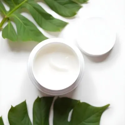 A White Jar With Cream And A Lid Surrounded By Two Green Leaves On A White Surface 43 Ed95