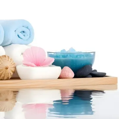 A Wooden Tray With Spa Items Including Blue Bath Salts In A Glass Bowl White And Pink Soaps A Small White Bottle And Decorative Shells There Are Also Two Rolled Towels In Light Blue And Pink The Background Is White 5 42c2