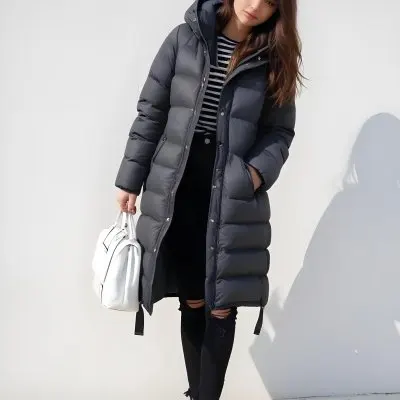 A Young Caucasian Woman With Long Brown Hair Wearing A Dark Gray Hooded Puffer Coat A Striped Shirt And Ripped Black Jeans She Is Holding A White Bag And Appears To Be Standing Outdoors In A Sunny Park 10 Cbe9