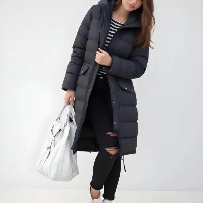 A Young Caucasian Woman With Long Brown Hair Wearing A Dark Gray Hooded Puffer Coat A Striped Shirt And Ripped Black Jeans She Is Holding A White Bag And Appears To Be Standing Outdoors In A Sunny Park 12 94d1