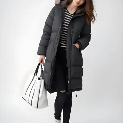 A Young Caucasian Woman With Long Brown Hair Wearing A Dark Gray Hooded Puffer Coat A Striped Shirt And Ripped Black Jeans She Is Holding A White Bag And Appears To Be Standing Outdoors In A Sunny Park 4 Ef9f