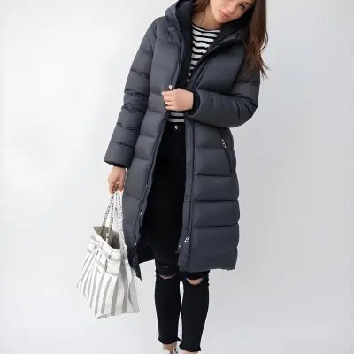 A Young Caucasian Woman With Long Brown Hair Wearing A Dark Gray Hooded Puffer Coat A Striped Shirt And Ripped Black Jeans She Is Holding A White Bag And Appears To Be Standing Outdoors In A Sunny Park 5 Cd08