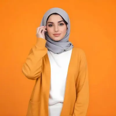 A Young Middle Eastern Woman With Dark Hair Wearing A Gray Hijab A White Sweater And A Orange Cardigan Standing Against A Orange Background 10 Debc