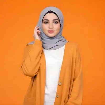 A Young Middle Eastern Woman With Dark Hair Wearing A Gray Hijab A White Sweater And A Orange Cardigan Standing Against A Orange Background 8 6a0e