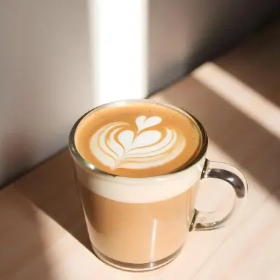 A Clear Glass Mug Filled With Latte Art Featuring A Heart Design On A Glass Table With A White Wall Background And Sunlight 11 Deda