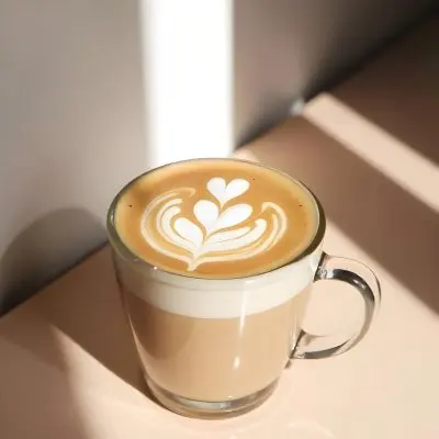 A Clear Glass Mug Filled With Latte Art Featuring A Heart Design On A Glass Table With A White Wall Background And Sunlight 25 B931