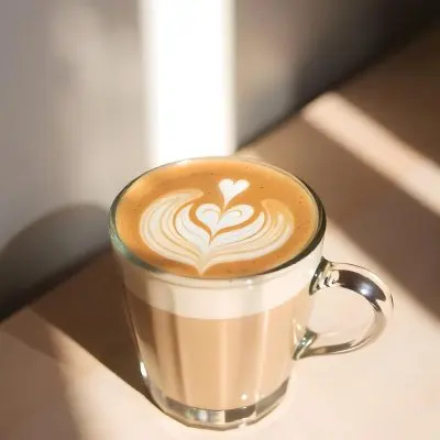 A Clear Glass Mug Filled With Latte Art Featuring A Heart Design On A Glass Table With A White Wall Background And Sunlight 3 5550