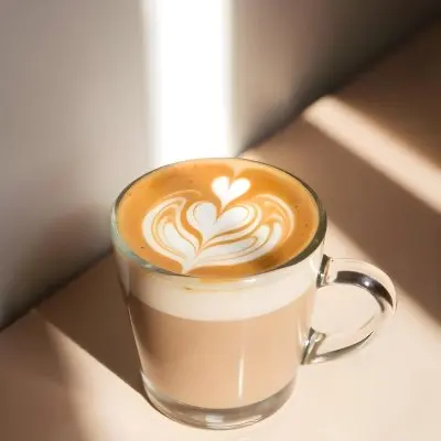 A Clear Glass Mug Filled With Latte Art Featuring A Heart Design On A Glass Table With A White Wall Background And Sunlight 40 77b2