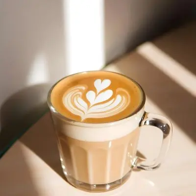 A Clear Glass Mug Filled With Latte Art Featuring A Heart Design On A Glass Table With A White Wall Background And Sunlight 42 9906